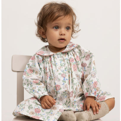 A young toddler with curly brown hair and blue eyes sits on a cream-colored wooden chair. The child is wearing a vintage-inspired long-sleeve floral dress with a Peter Pan collar, adorned with pink and blue flowers on a white background. Small white buttons run down the front. The baby also wears beige knitted socks with lace trim. The neutral background and soft lighting enhance the delicate and timeless aesthetic of the outfit