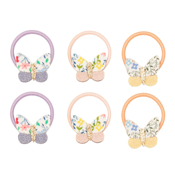 A set of six butterfly-themed mini hair ties from Sonny Bear Mimi & Lula. Each hair tie features a small butterfly decoration with fabric wings in floral and pastel patterns, along with glittery gold accents in the center. The elastic bands come in soft shades of lilac, peach, and cream, complementing the delicate butterfly design